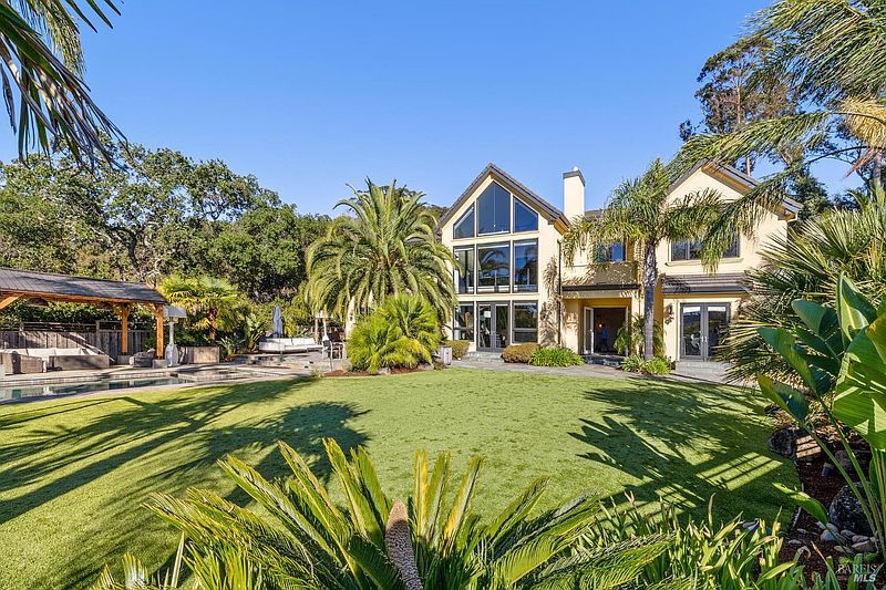 This image showcases the expansive backyard of a luxurious home, featuring a well-manicured lawn, mature palm trees, and a covered outdoor living area with a pool. The house itself boasts large windows and a light-colored exterior, creating an inviting and upscale atmosphere perfect for entertaining or relaxing.