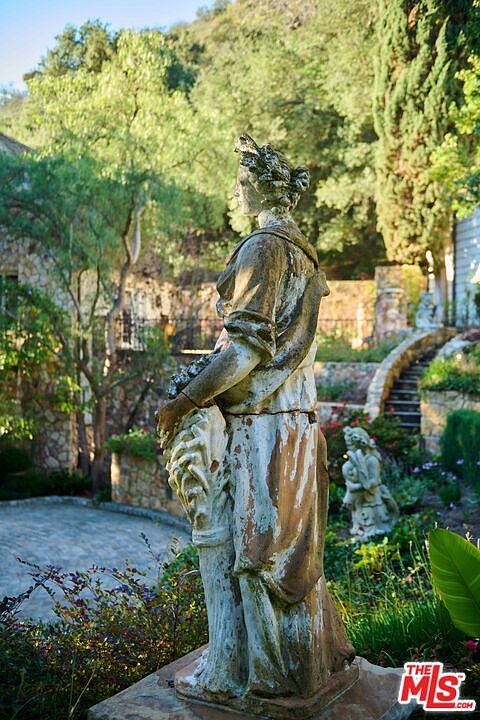 A weathered stone statue of a woman stands prominently in the foreground of a lush, terraced garden. The background features a stone-walled property nestled against a hillside with mature trees and a winding stone staircase. The scene evokes a classic, European-inspired estate atmosphere with a focus on landscape design and architectural detail.