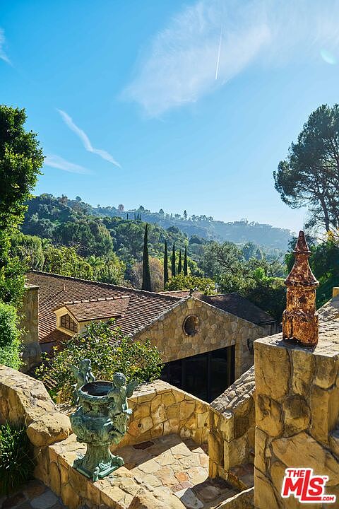 This elevated perspective captures a charming stone-clad residence nestled within a lush, hilly landscape. The foreground features a decorative stone terrace adorned with a classic cherub-themed urn, leading the eye toward the home's rustic tile roof and the expansive, verdant mountain backdrop. The scene evokes a serene, Mediterranean-inspired atmosphere, highlighting the property's integration with its natural surroundings.