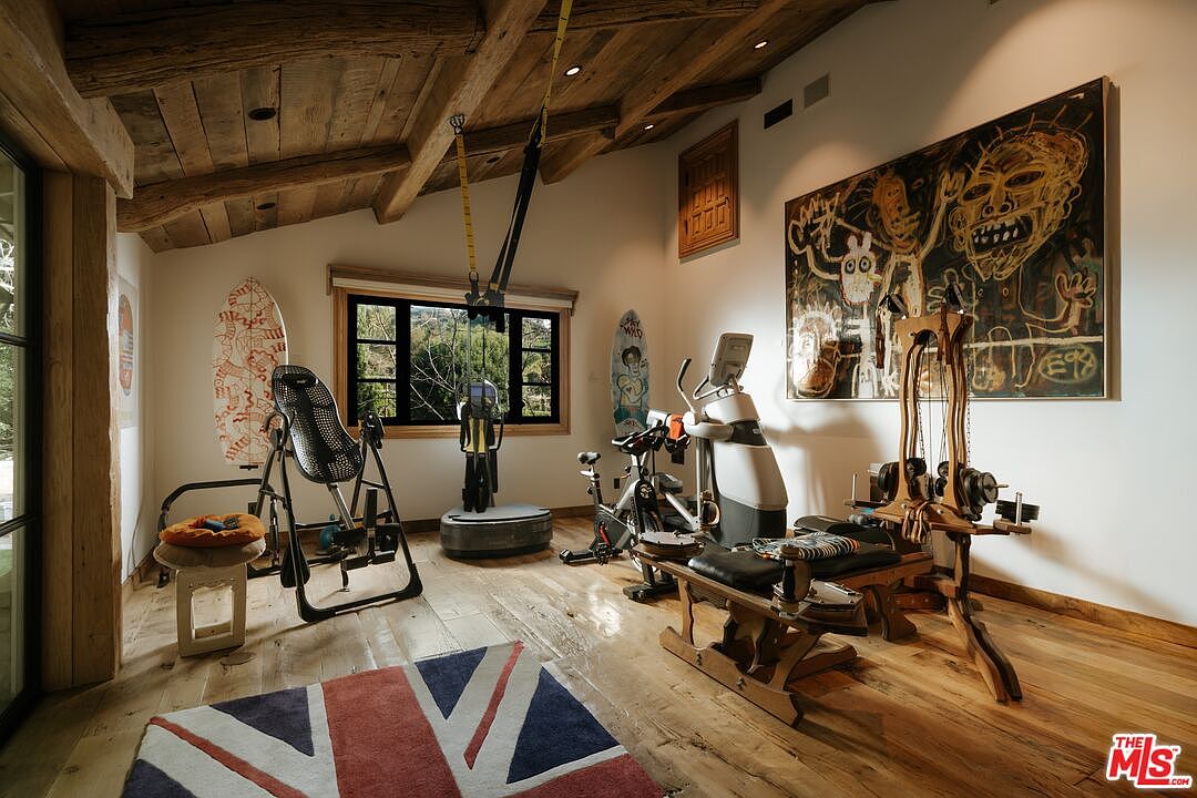 This rustic-style home gym features exposed wooden ceiling beams, hardwood flooring, and a large, vibrant abstract painting on the wall. The room is well-equipped with various fitness machines, including a stationary bike, a vibration platform, and a weight training station, all set against a backdrop of natural light from the window. A Union Jack rug adds a pop of color to the space, creating an energetic and personalized workout environment.