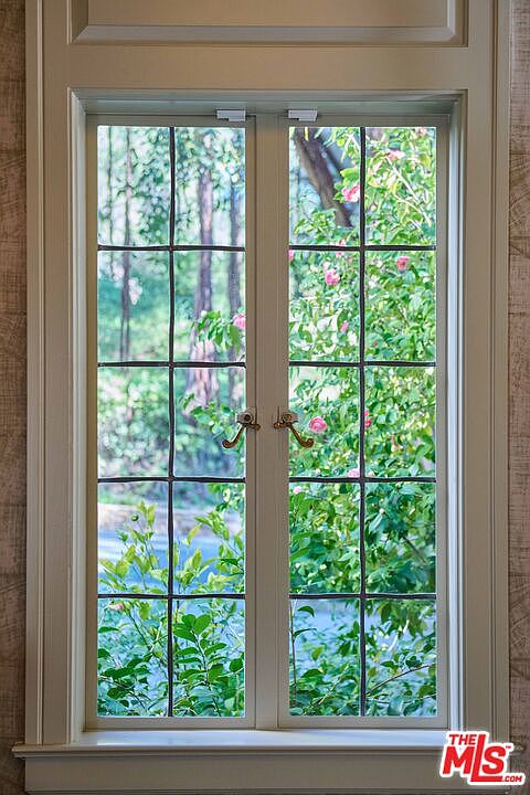 This image captures a classic, double-casement window with a grid-patterned muntin design, framed by elegant, light-colored woodwork. The window provides a clear, serene view of lush green foliage and blooming pink flowers outside, creating a peaceful and inviting atmosphere. The perspective is a direct, eye-level shot that emphasizes the craftsmanship of the window frame and the natural light filtering into the interior space.