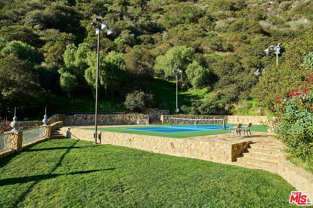 This image showcases a private tennis court nestled against a lush, tree-covered hillside, creating a serene and secluded recreational space. The court features a vibrant blue and green surface, complemented by stone retaining walls and a well-manicured lawn in the foreground. The perspective is a wide, elevated shot that captures the integration of the sports facility into the natural landscape, emphasizing the property's expansive outdoor amenities.