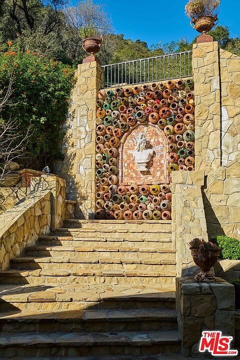This striking outdoor staircase features a Mediterranean-inspired stone wall adorned with a collection of terracotta pots and a central classical bust sculpture. The scene is framed by lush greenery and stone masonry, creating a dramatic and artistic focal point for a garden or landscape design. The perspective is a low-angle, eye-level shot that emphasizes the texture of the stone and the intricate arrangement of the pottery.
