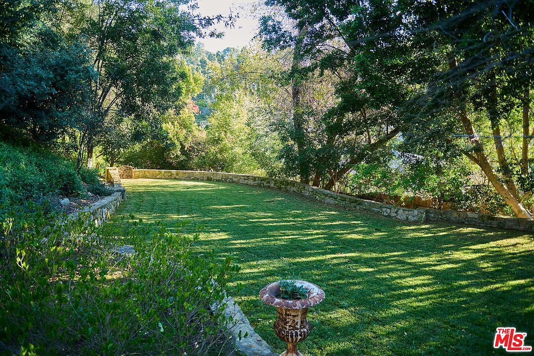This serene backyard features a long, manicured lawn bordered by a rustic stone retaining wall and lush, mature trees. A decorative stone urn sits in the foreground, adding a touch of classic elegance to the peaceful, shaded landscape. The perspective is a wide, eye-level shot that captures the depth and tranquil atmosphere of the private outdoor space.