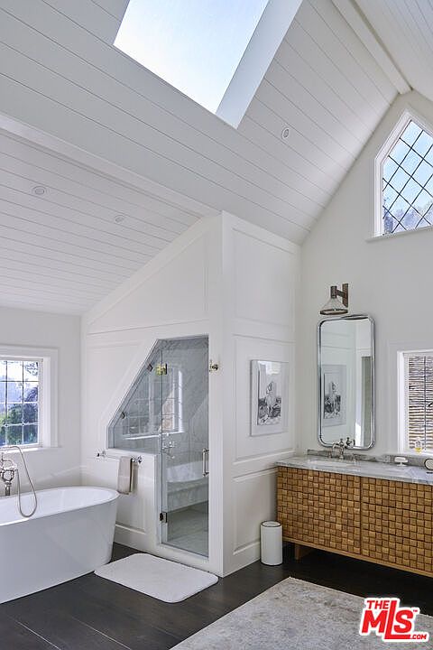 This bright and airy primary bathroom features a vaulted ceiling with a large skylight, creating a spacious and light-filled atmosphere. The room includes a sleek freestanding soaking tub, a custom glass-enclosed shower with unique angled architecture, and a vanity with a textured, wood-block facade. The combination of white shiplap walls, dark flooring, and diamond-paned windows gives the space a sophisticated, modern farmhouse aesthetic.