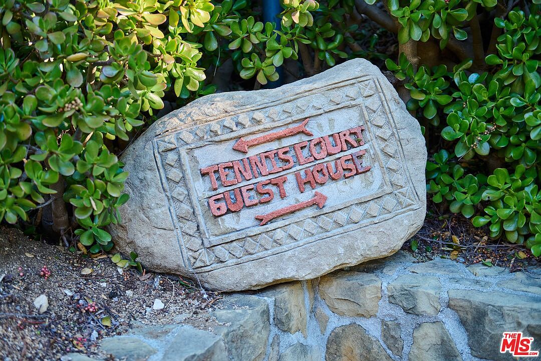 A close-up, eye-level shot of a decorative stone sign nestled within a lush garden setting. The carved rock features directional arrows and the text 'TENNIS COURT' and 'GUEST HOUSE', set against a backdrop of vibrant green jade plants and a rustic stone retaining wall. The scene conveys a sense of well-maintained, upscale landscaping and clear property navigation.
