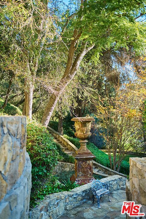 This serene garden scene features a classic stone-paved pathway winding through lush, mature landscaping and tiered stone retaining walls. A prominent, ornate antique-style urn sits as a focal point, complemented by a small, decorative metal garden chair. The perspective is a low-angle, eye-level shot that emphasizes the textured stonework and the dappled sunlight filtering through the dense canopy of trees.