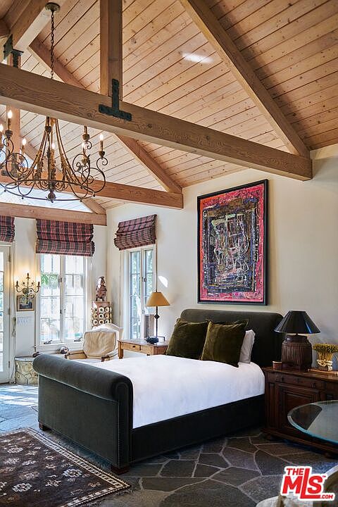 This primary bedroom features a rustic, vaulted ceiling with exposed wooden beams and a large, ornate iron chandelier. The room is anchored by a dark upholstered sleigh bed, complemented by a vibrant, abstract piece of art on the wall and a stone-tiled floor. The space exudes a cozy, lodge-like atmosphere with warm lighting and classic window treatments.