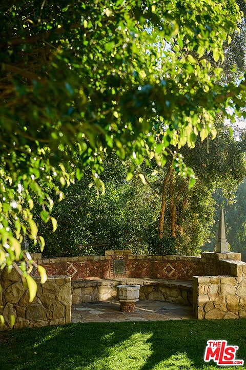 This serene garden setting features a curved, stone-built bench integrated into a low retaining wall, accented with decorative tile inlays. A small, ornate stone pedestal sits in the center of the flagstone patio, framed by lush, overhanging tree branches that create a dappled light effect. The scene evokes a peaceful, Mediterranean-inspired retreat perfect for quiet outdoor relaxation.