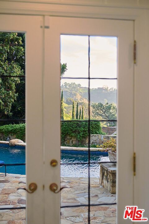 This view is captured from inside looking out through a set of classic white French doors, framing a serene backyard scene. The perspective highlights a sparkling blue swimming pool set against a stone patio, with lush greenery and rolling hills visible in the background. The composition creates a seamless transition between the interior living space and the tranquil outdoor oasis.