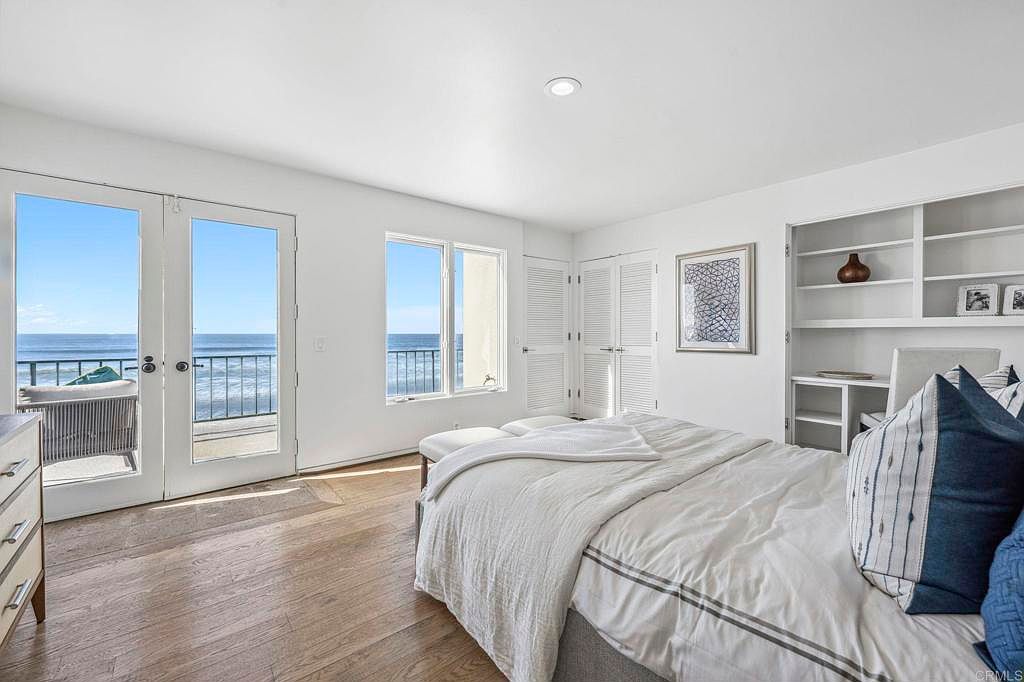 This is a bright and airy primary bedroom with a view of the ocean. The room features hardwood floors, white walls, and large windows and doors that let in plenty of natural light. A comfortable bed with white linens and blue accent pillows is the focal point, complemented by built-in shelving and a serene atmosphere.
