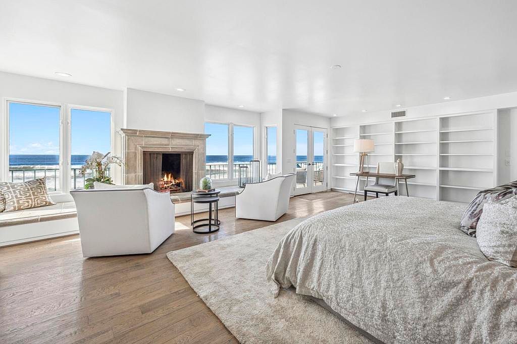 This is a luxurious primary bedroom featuring a fireplace, built-in bookshelves, and large windows with ocean views. The room is spacious and well-lit, with hardwood floors and a neutral color palette creating a serene atmosphere. The bed is prominently featured, suggesting comfort and relaxation.