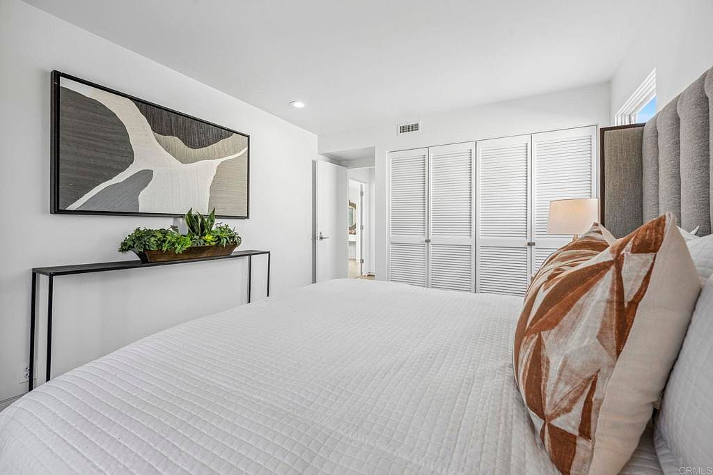 This is a bright and airy primary bedroom featuring a large bed with white quilted bedding and decorative pillows. A modern console table with greenery sits beneath an abstract art piece, adding a touch of sophistication. The room is painted in a clean white, enhancing the sense of space and tranquility, with closet doors lining the back wall.