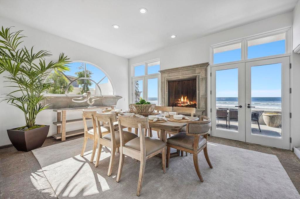 This is an interior shot of a dining room featuring a wooden dining table set for a meal, surrounded by six chairs. A fireplace is visible in the background, and large glass doors offer a view of the ocean. The room is well-lit with natural light, creating a bright and inviting atmosphere.