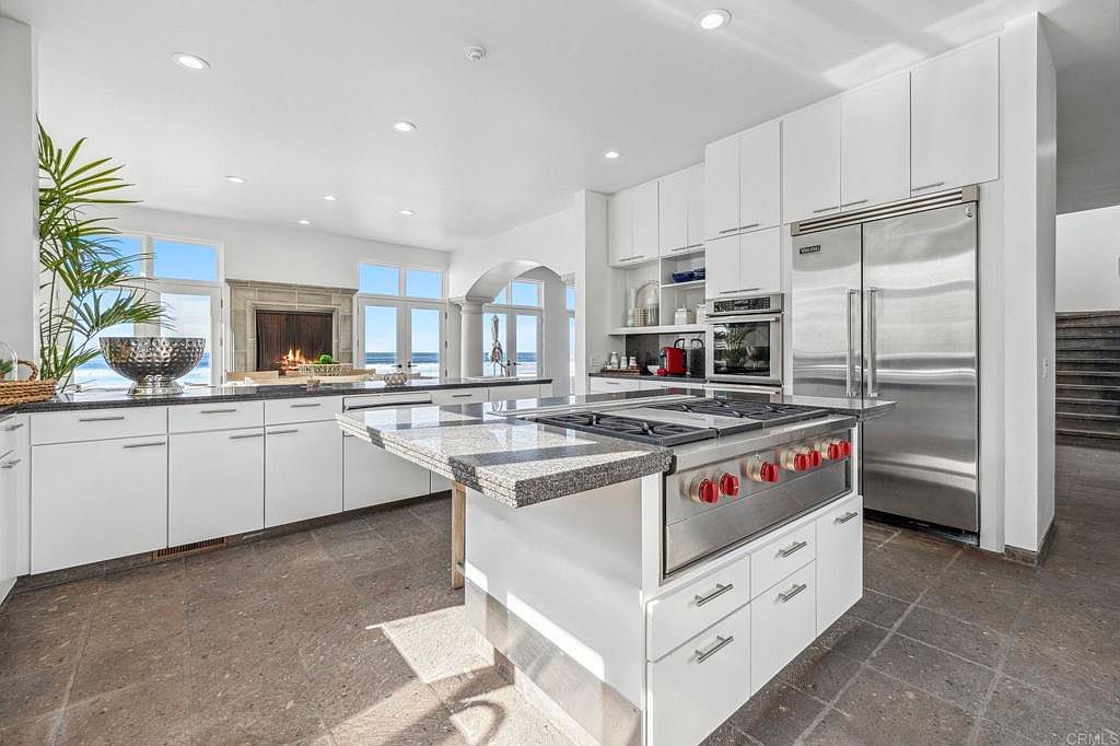 This is a bright, modern kitchen featuring white cabinetry, stainless steel appliances, and a large island with a granite countertop. The kitchen includes a professional-grade range with red knobs and a stainless steel refrigerator. Large windows offer a view of the ocean, and the flooring appears to be stone tile, creating a luxurious and functional space.