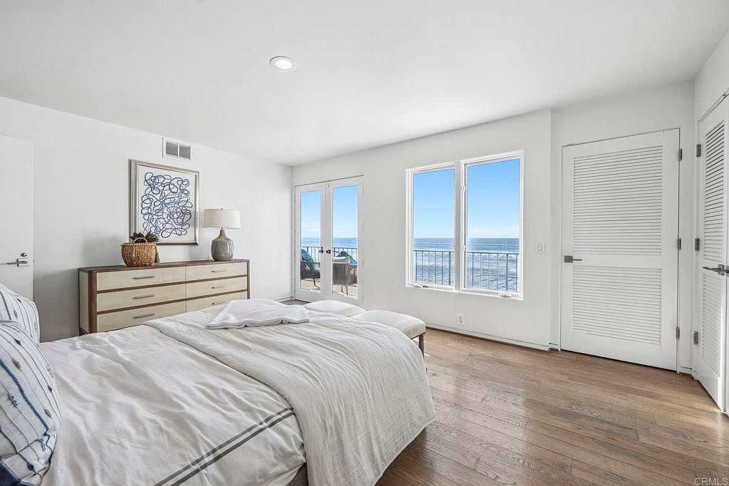 This is a bright and airy primary bedroom featuring hardwood floors, white walls, and large windows with ocean views. The room includes a dresser with a lamp and artwork, a bed with white linens, and access to a balcony through French doors. The overall impression is clean, modern, and serene.