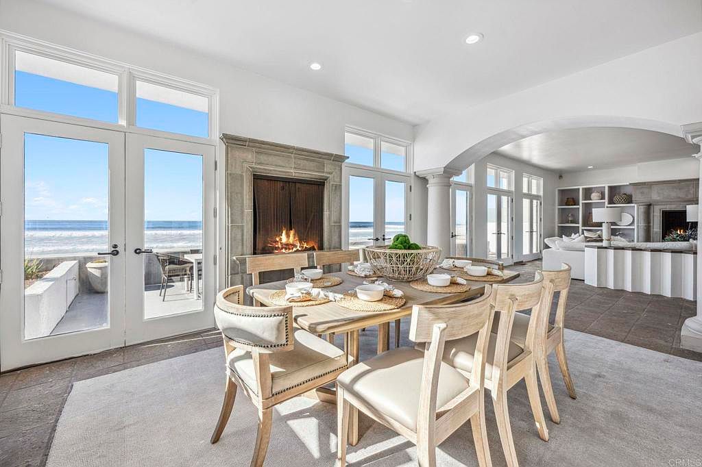 This is an interior shot of a dining room with a beach view. The dining area features a wooden table set for a meal, surrounded by upholstered chairs. A fireplace adds warmth, while large windows and doors offer an ocean view, creating a luxurious and inviting atmosphere.