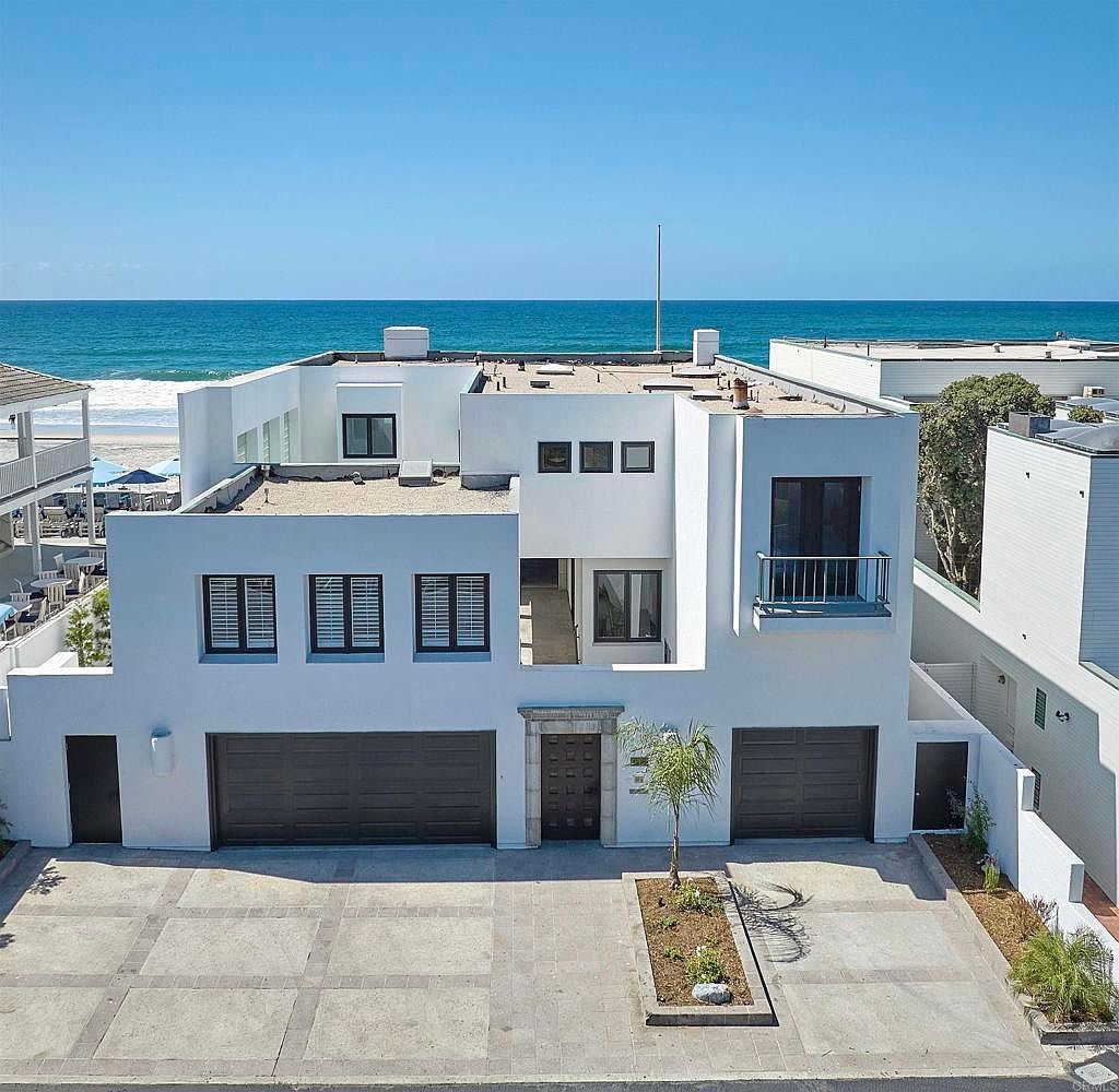 This is a front exterior view of a modern, white beach house. The house features a flat roof, dark-framed windows, and two garage doors. A small palm tree is planted in the front yard, and the ocean is visible in the background, suggesting a prime coastal location.