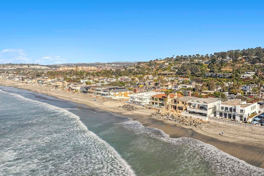 This aerial view showcases a stunning beachfront property with multiple buildings along the coastline. The architecture is modern and luxurious, with clean lines and large windows. The scene captures the beauty of coastal living, with the ocean waves gently meeting the sandy beach, and the clear blue sky above.