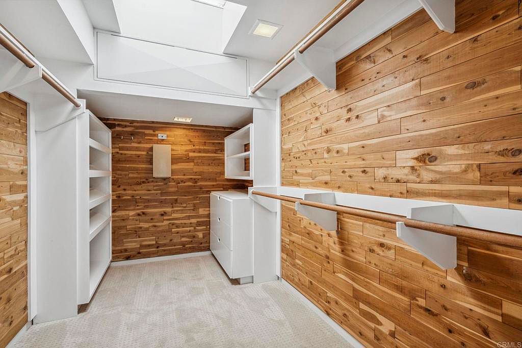 This is a well-organized walk-in closet featuring wooden plank walls, white shelving units, and wooden closet rods. The closet is illuminated by a skylight and recessed lighting, creating a bright and inviting space. The overall impression is one of luxury and functionality.