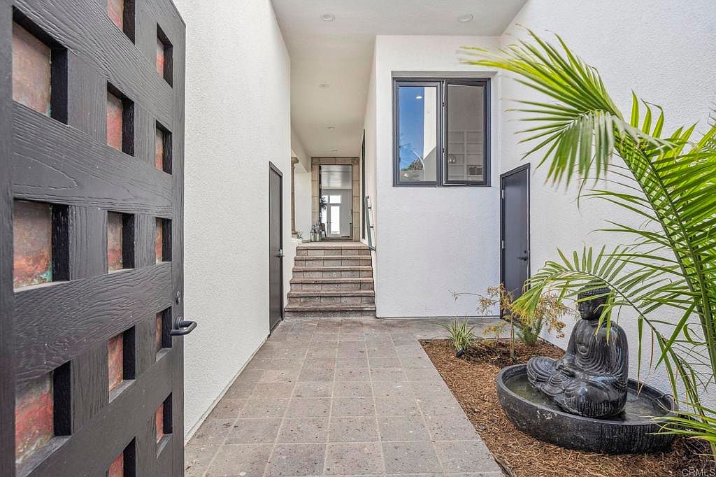 This image showcases an inviting entryway with a modern aesthetic. A unique, dark-toned door with square cutouts is partially open, leading into a tiled courtyard featuring a Buddha statue water feature and a set of stairs leading to the main entrance. The white walls and black-framed window add to the contemporary feel, creating a serene and stylish welcome.