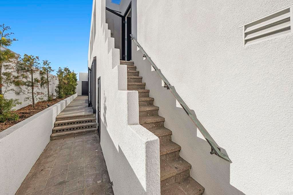 This exterior side view showcases a modern building with clean architectural lines and a textured white facade. A staircase with a metal handrail leads to an upper level, while a lower walkway is bordered by a low wall and landscaping. The image highlights the building's design and outdoor access.