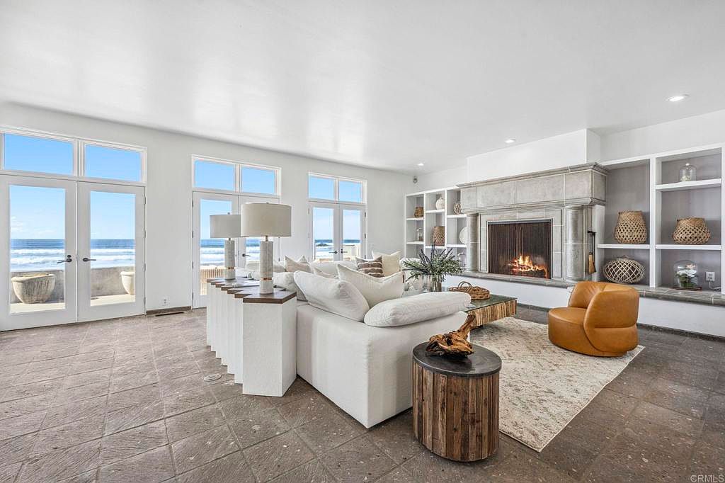 This is an interior shot of a luxurious living room featuring a large white sectional sofa, a stone fireplace with built-in shelving, and floor-to-ceiling windows offering ocean views. The room is decorated in a neutral color palette with natural wood accents, creating a serene and upscale atmosphere. The flooring is a large format tile.