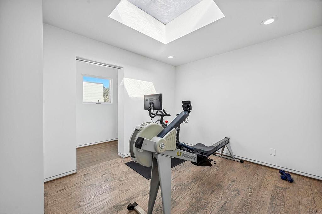 This is an interior shot of a home gym featuring a Concept2 rowing machine as the focal point. The room is well-lit with a skylight and recessed lighting, and the hardwood floors add a touch of warmth. A window provides natural light and a view to the outside, enhancing the workout environment.