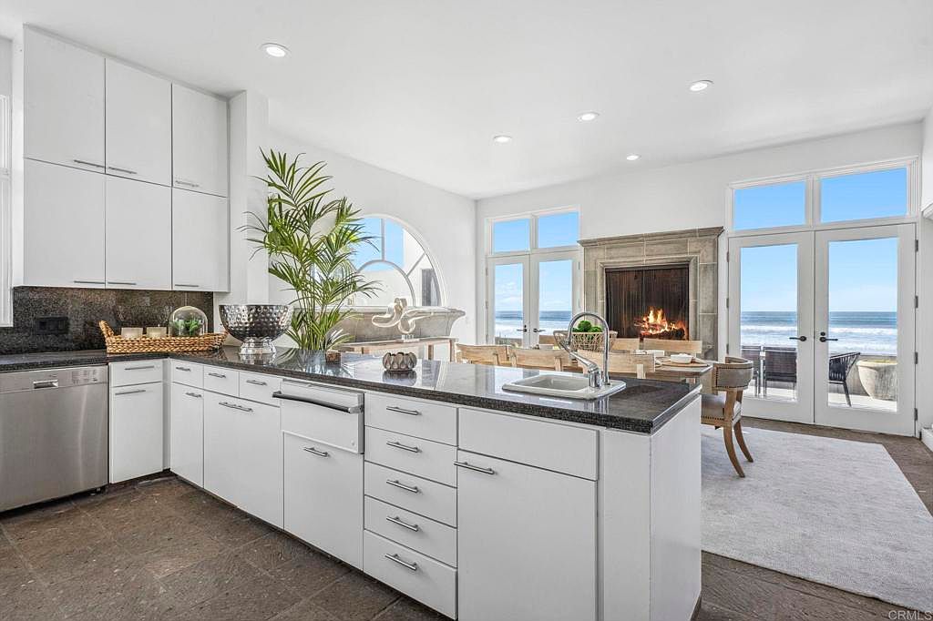 This is a bright and airy kitchen with white cabinets, dark countertops, and stainless steel appliances. The kitchen island features a sink and ample storage. Large windows and doors offer stunning ocean views, creating a seamless indoor-outdoor living experience.