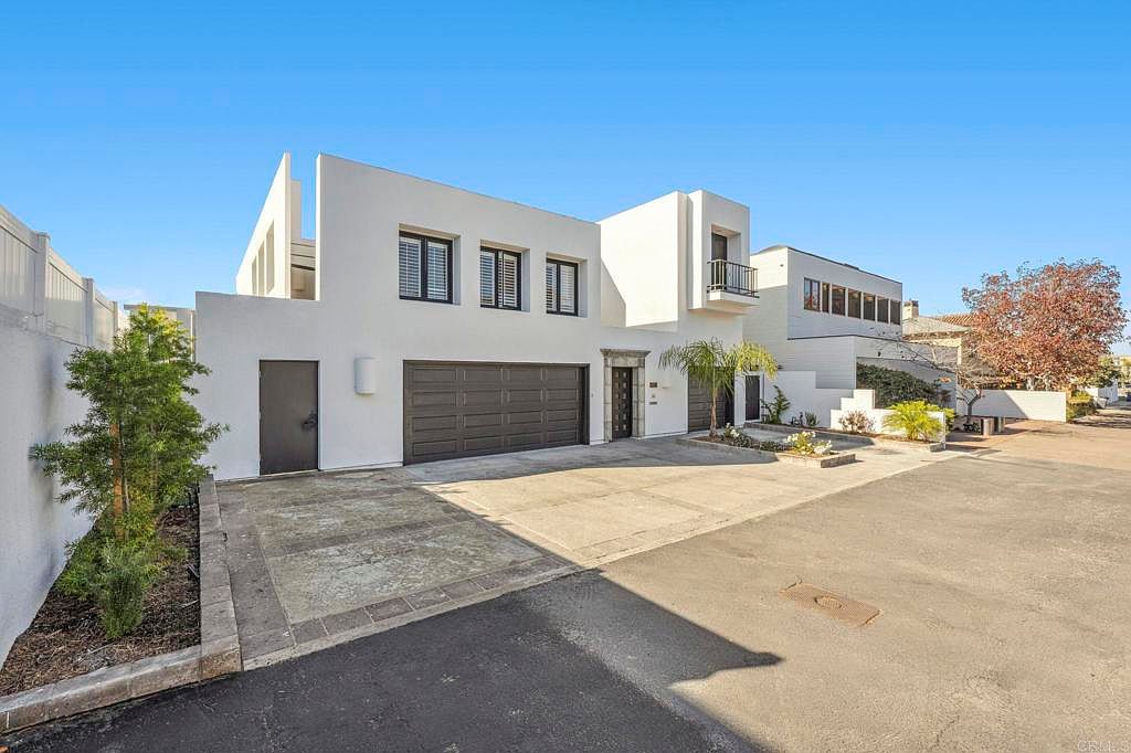 This is a front exterior view of a modern two-story home with a white facade and dark-framed windows. The property features a two-car garage, a separate entrance door, and a small balcony on the second floor. The driveway and landscaping add to the curb appeal, presenting a clean and contemporary aesthetic.