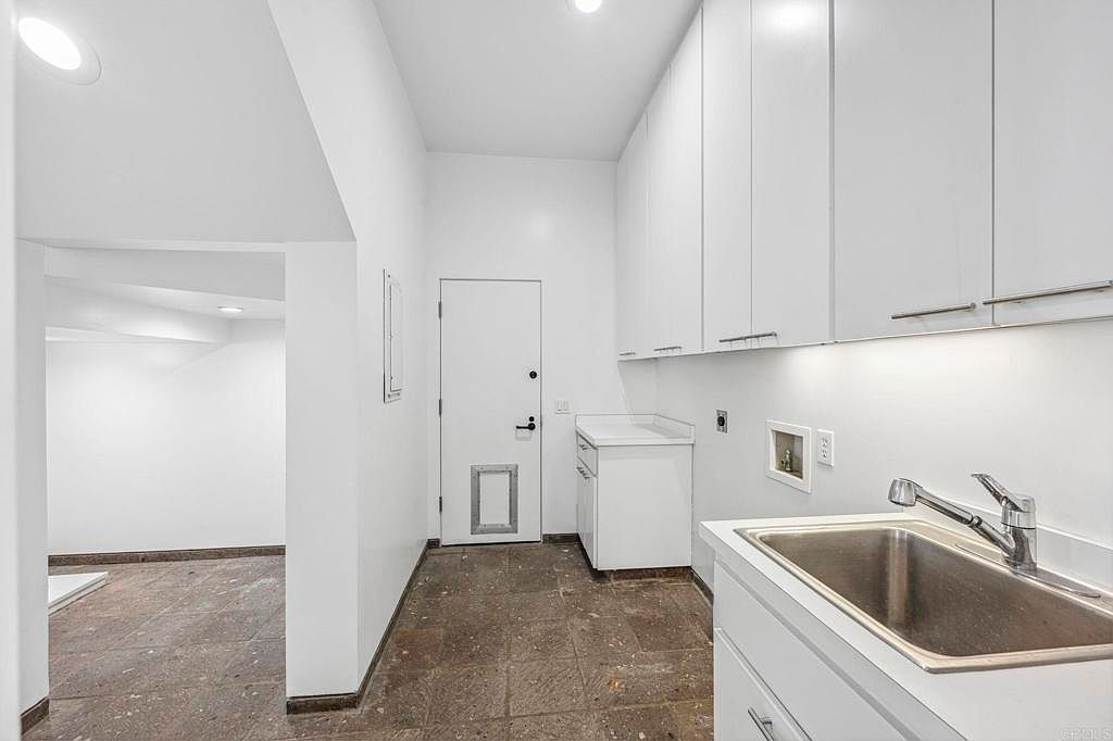 This is an interior shot of a laundry room featuring white cabinetry and a stainless steel sink. The room has a clean and modern aesthetic, with a door leading to another room. The flooring appears to be tile, and the overall impression is one of functionality and organization.