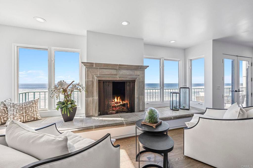 This is an interior shot of a luxurious living room featuring a stone fireplace with a burning fire. Large windows offer stunning ocean views, and the room is furnished with elegant white armchairs and a dark wood floor. The overall impression is one of sophisticated comfort and coastal living.