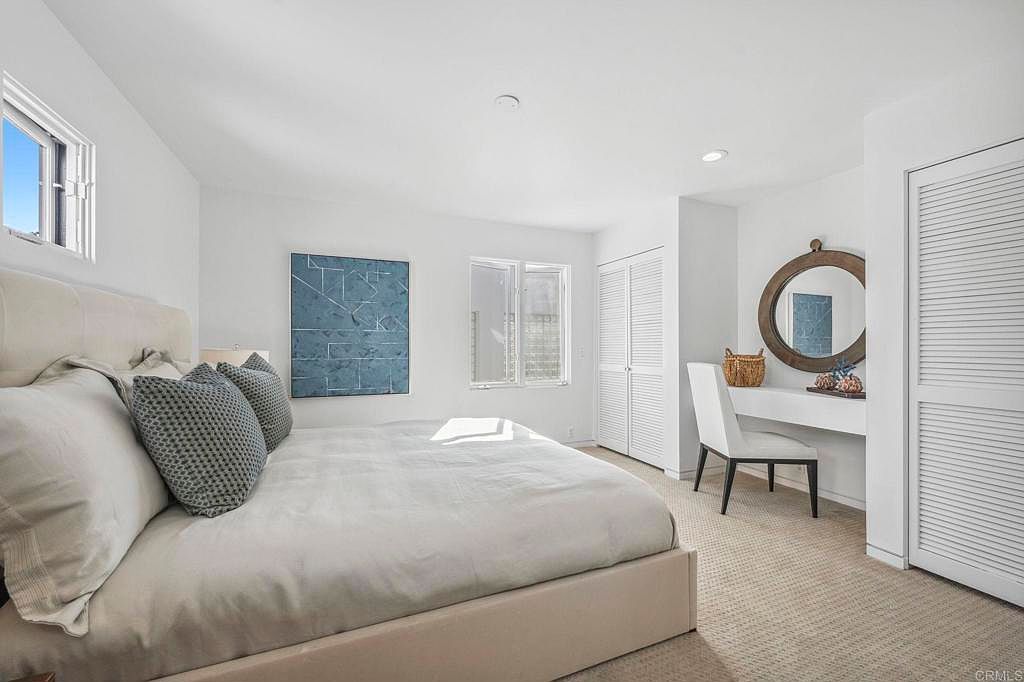 This is a bright and airy primary bedroom featuring a large bed with white linens and decorative pillows. The room is painted in a clean white, complemented by a blue abstract artwork and natural light streaming through the windows. A built-in vanity with a round mirror and a white chair adds a touch of elegance, while the carpeted floor provides warmth and comfort.