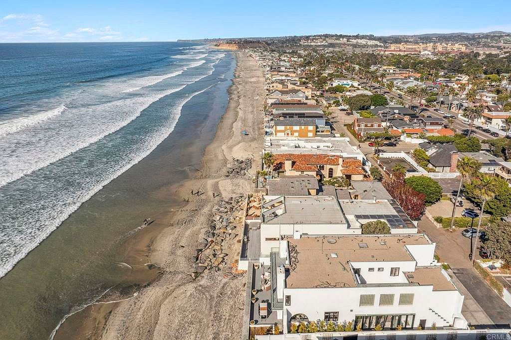 This aerial view showcases a stunning beachfront property with direct access to the sandy shore and ocean. The homes feature a mix of architectural styles, with some having rooftop patios and decks. The image captures the proximity to the beach and the overall coastal lifestyle.