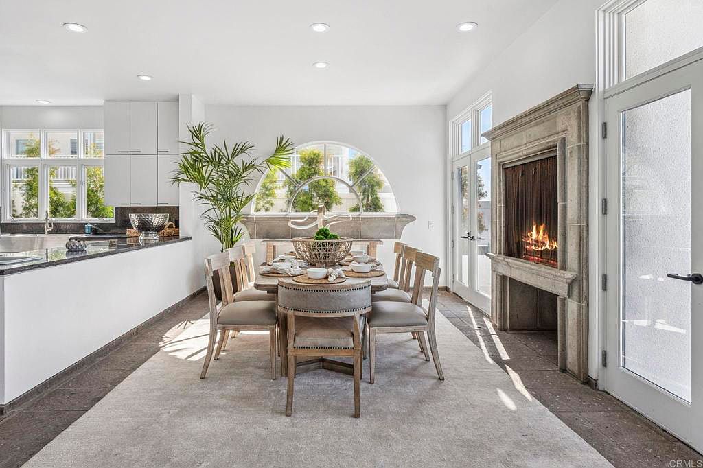 This is an interior shot of a dining room featuring a round dining table set for a meal, surrounded by upholstered chairs. A large arched window provides natural light, and a fireplace adds a cozy ambiance. The room is decorated in neutral tones with a modern aesthetic, creating an inviting space for dining and entertaining.