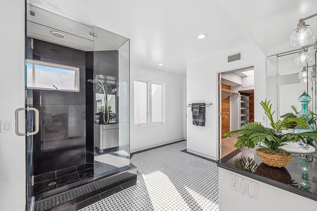 This is a bright and modern primary bathroom featuring a glass-enclosed shower with black tile, a double window providing natural light, and a black and white patterned tile floor. The vanity area includes a dark countertop, decorative plants, and mirrored wall, creating a clean and stylish space. A walk-in closet is visible through a doorway.