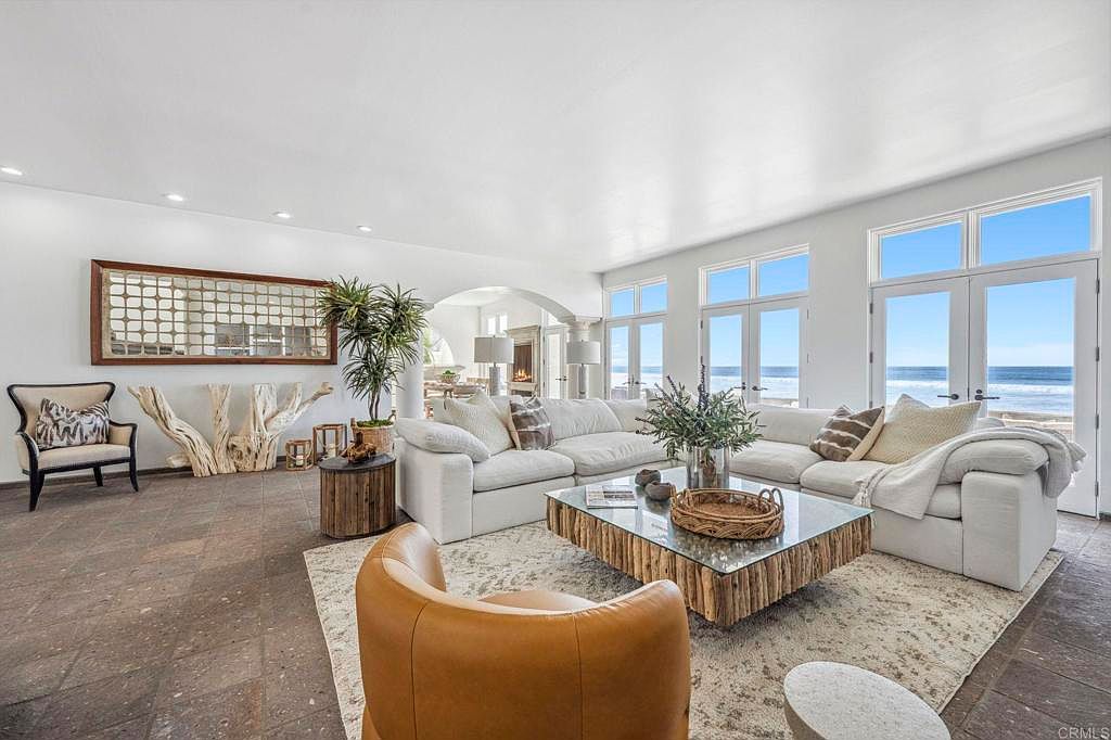 This is a bright and airy living room with a coastal aesthetic. The room features a large white sectional sofa, a unique wooden coffee table with a glass top, and a decorative mirror on the wall. Large windows offer ocean views, enhancing the room's serene and luxurious atmosphere.
