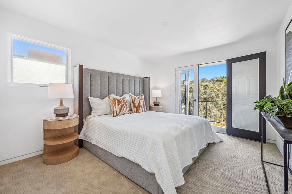 This is a bright and airy primary bedroom featuring a large bed with a gray upholstered headboard and white bedding accented with patterned pillows. Natural light floods the room through a window and a glass door leading to a balcony, enhancing the serene atmosphere. The room is decorated in a modern style with neutral tones and minimalist furniture.