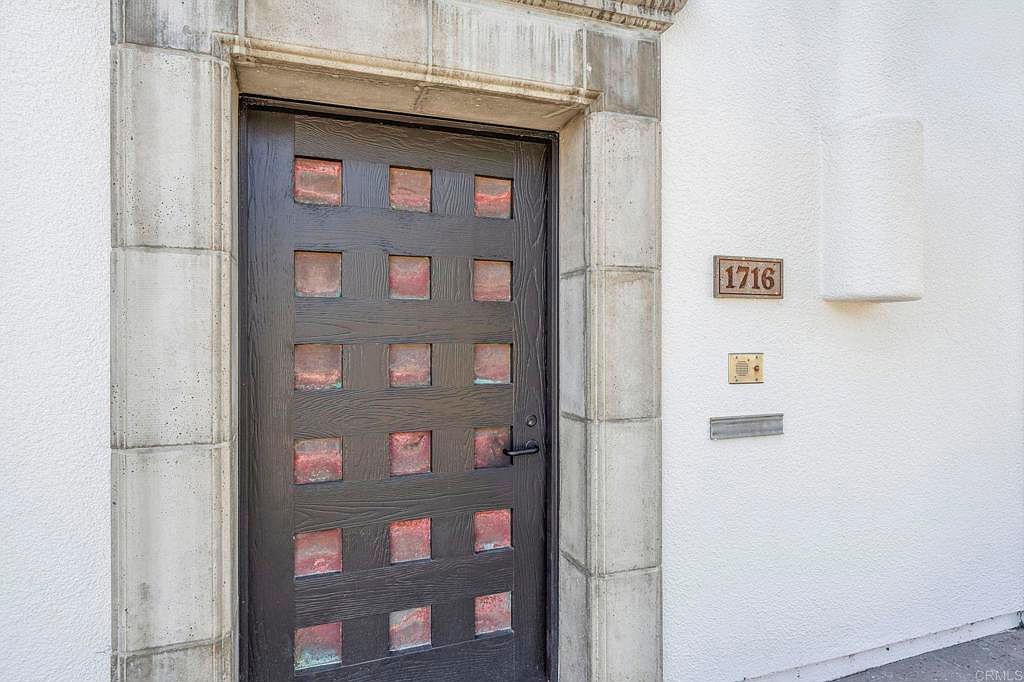 This image showcases an elegant entryway featuring a dark wood door with square glass inserts, framed by a light-colored stone surround. The white stucco wall adjacent to the door includes the house number '1716', a doorbell, and a mail slot. The perspective is a medium shot, highlighting the architectural details and creating a welcoming impression.