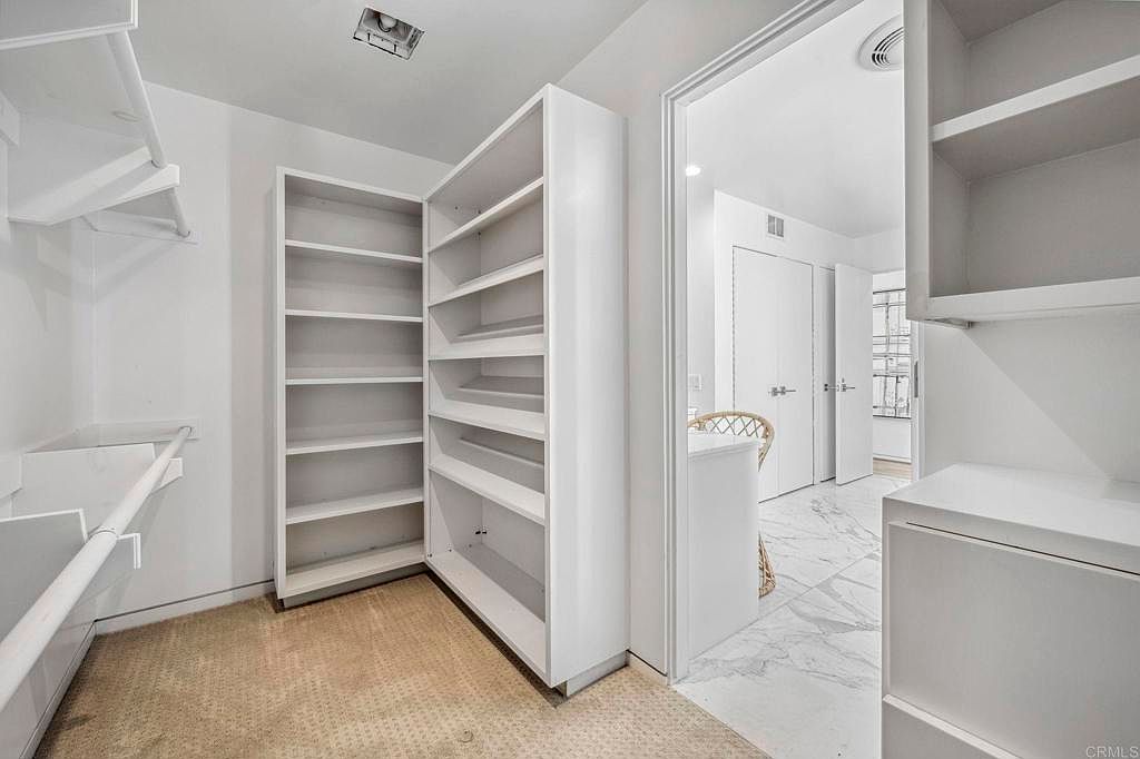 This is a well-organized walk-in closet featuring built-in shelving and storage units. The closet has a neutral color palette with white shelves and beige carpet. The space is bright and appears spacious, offering ample storage for clothing and accessories.