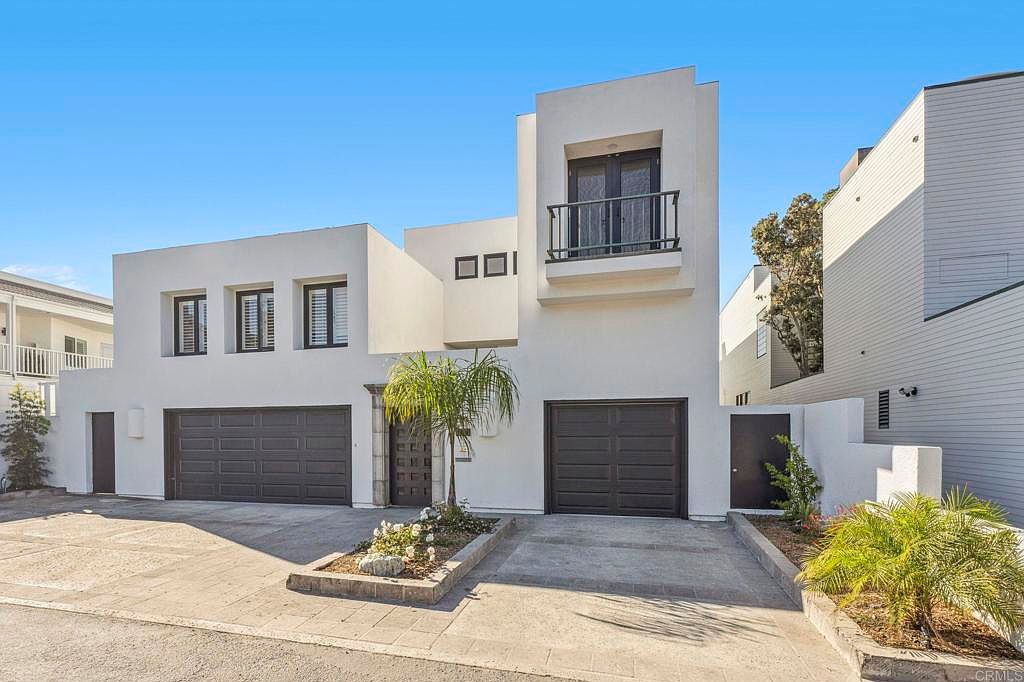 This is a front exterior view of a modern, two-story home with a white facade. The house features two garage doors, a small balcony, and well-maintained landscaping with palm trees and flowering plants. The architectural style is contemporary, emphasizing clean lines and geometric shapes.