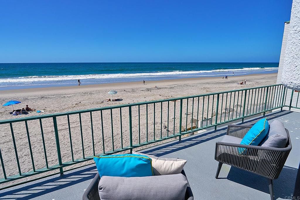 This image showcases a serene balcony view overlooking a sandy beach and ocean. The balcony features a green railing and comfortable outdoor seating with stylish cushions. The scene evokes a sense of relaxation and coastal living, highlighting the property's desirable location and outdoor space.