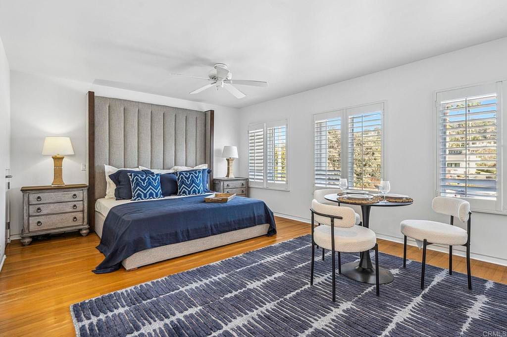 This is a primary bedroom featuring a king-size bed with a tall, upholstered headboard and blue bedding. The room has hardwood floors covered by a blue and white patterned rug, and there is a small dining table with chairs near the windows. The style is modern and comfortable, creating a relaxing atmosphere.