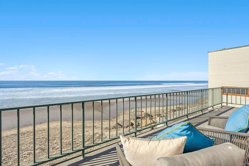 This image showcases a beautiful patio or balcony overlooking a sandy beach and the ocean. The space is furnished with comfortable seating, including pillows, suggesting a relaxing outdoor living area. A railing provides safety and enhances the view, making it an ideal spot for enjoying the coastal scenery.