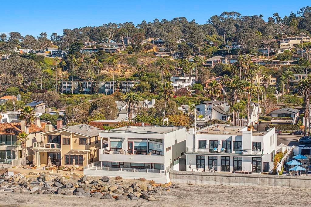 This image showcases the front exterior of three modern beachfront homes. The houses feature a mix of architectural styles, with one having a traditional tan facade and the other two boasting clean, white, contemporary designs. Large windows and balconies offer stunning ocean views, while the rocky shoreline adds to the property's appeal.