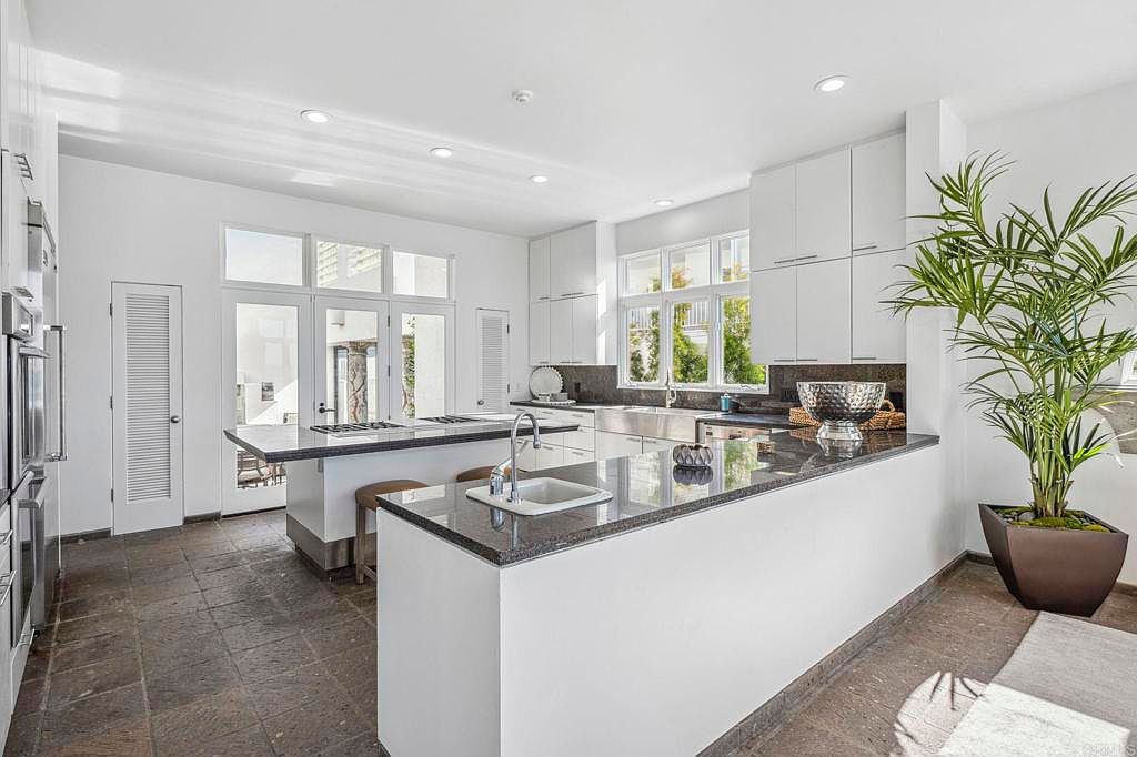 This is a bright and spacious kitchen featuring white cabinetry, granite countertops, and stainless steel appliances. A large island provides ample workspace and seating, while large windows offer natural light. The flooring appears to be a dark tile, adding contrast to the light-colored cabinetry.