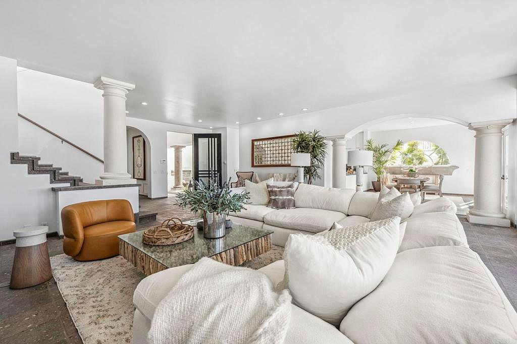 This is a spacious living room featuring a large, comfortable white sectional sofa, a unique glass-topped coffee table with a wooden base, and a tan leather chair. The room is bright and airy, with white walls and architectural columns adding a touch of elegance. The flooring appears to be stone, and the overall impression is one of relaxed luxury.