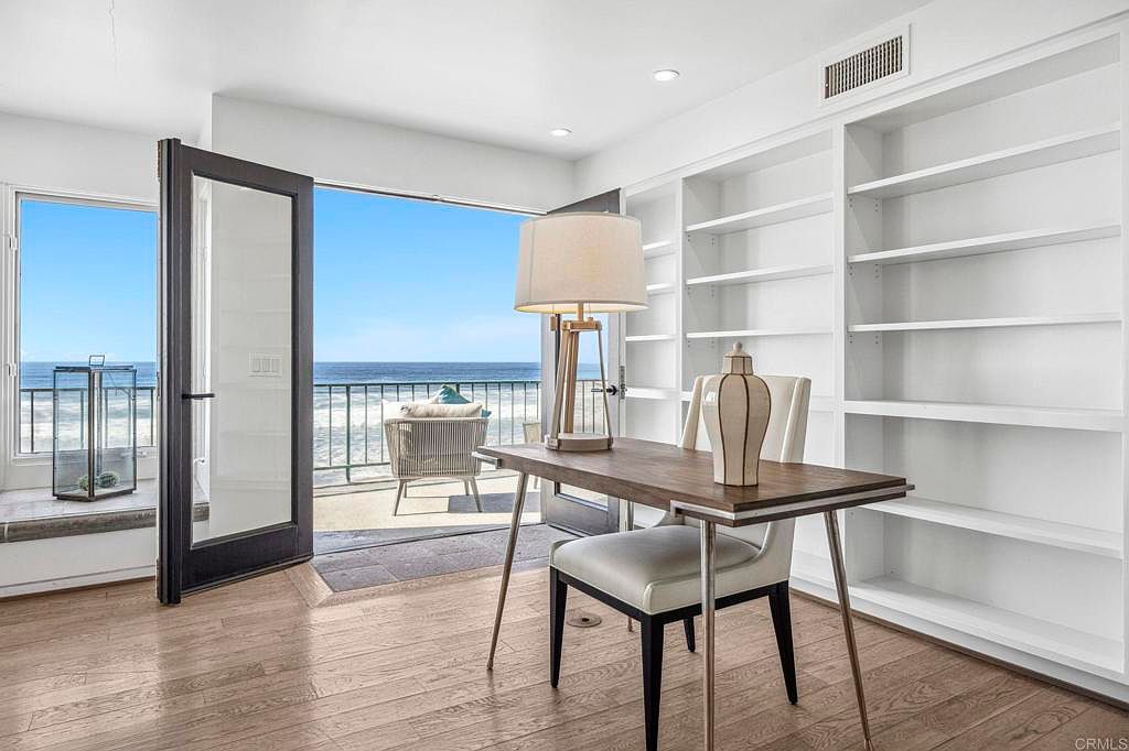 This interior shot showcases a bright and airy office space with a stunning ocean view. The room features a wooden desk with a white chair, built-in white bookshelves, and large glass doors leading to a balcony. The natural light and minimalist design create a serene and productive atmosphere, perfect for a home office.