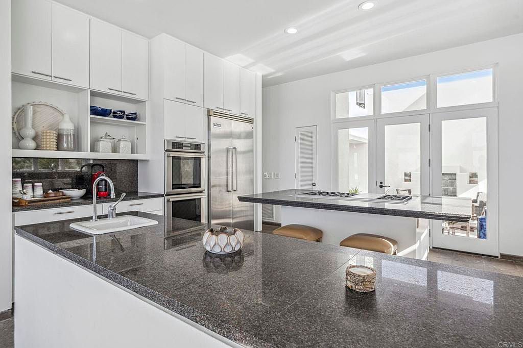 This is a bright and modern kitchen featuring white cabinetry, stainless steel appliances, and a large dark countertop island with seating. The kitchen is well-lit with natural light coming through the large windows and doors, creating an open and airy feel. The perspective is from the front of the island, showcasing the sink and faucet, as well as the decorative items on the countertop.
