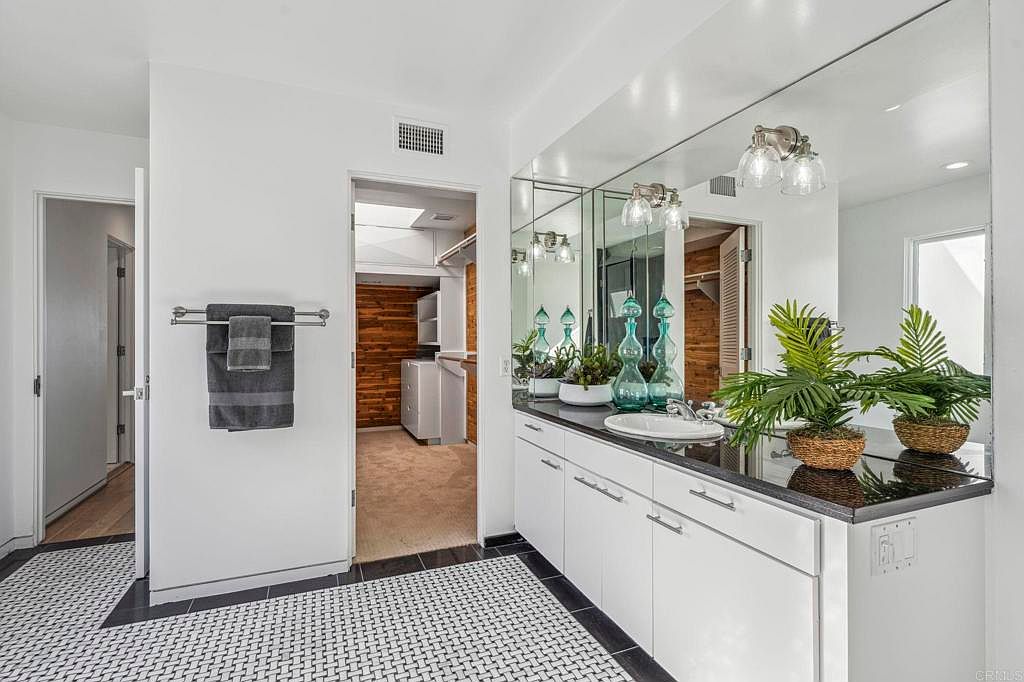 This bathroom features a white vanity with a black countertop, a large mirror with decorative lighting, and a black and white patterned tile floor. The room has a clean and modern aesthetic, with a glimpse into an adjacent laundry area with wood paneling. The perspective is from the doorway, showcasing the vanity and the adjacent room.